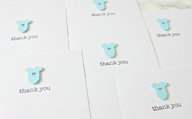 May include: Set of five thank you cards with a light blue baby onesie cutout with a small heart in the center. Each card says "thank you" in black text.