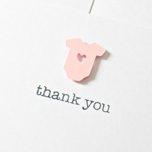 May include: A pink paper baby onesie with a heart cut out, with the text "thank you" printed below.
