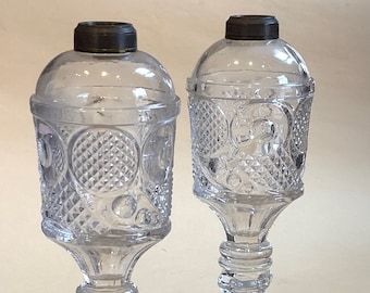 May include: Two clear glass oil lamps with a cut glass design. The lamps have a round base and a hexagonal base.