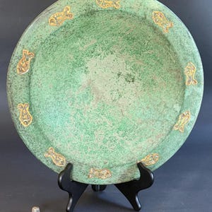 May include: A round, green plate with gold-colored fish designs around the rim. The plate has a distressed, aged appearance and is displayed on a black stand. A small, silver thimble is in the foreground.