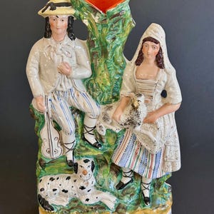 May include: A vintage ceramic figurine of a couple with a dog and a sheep. The man is wearing a hat and holding a violin. The woman is holding a sheep. The figurine is painted in bright colors and has a green tree trunk in the center.