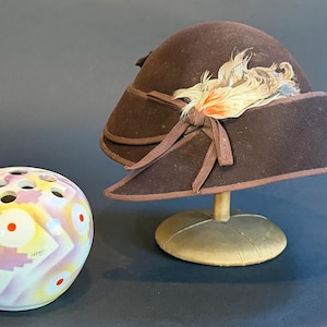 May include: A brown felt hat with a decorative feather and bow, displayed on a hat stand, next to a round, multi-colored vase with several holes. The vase has a geometric design with red and yellow accents.