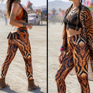 May include: Two women wearing matching tiger print leggings and tops. One woman wears a fringed orange top and a brown belt with a pouch. The other woman wears a black mesh top and a matching tiger print kimono. The brand name "PSYTE CLOTHING" is visible.