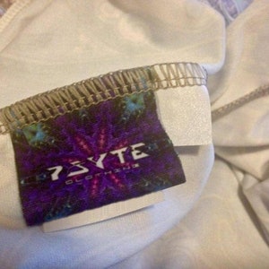 Pu&ograve; includere: Primo piano di un'etichetta di abbigliamento con la scritta "PSYTE CLOTHING" in bianco su sfondo viola. L'etichetta presenta un design psichedelico colorato ed &egrave; cucita su tessuto bianco.