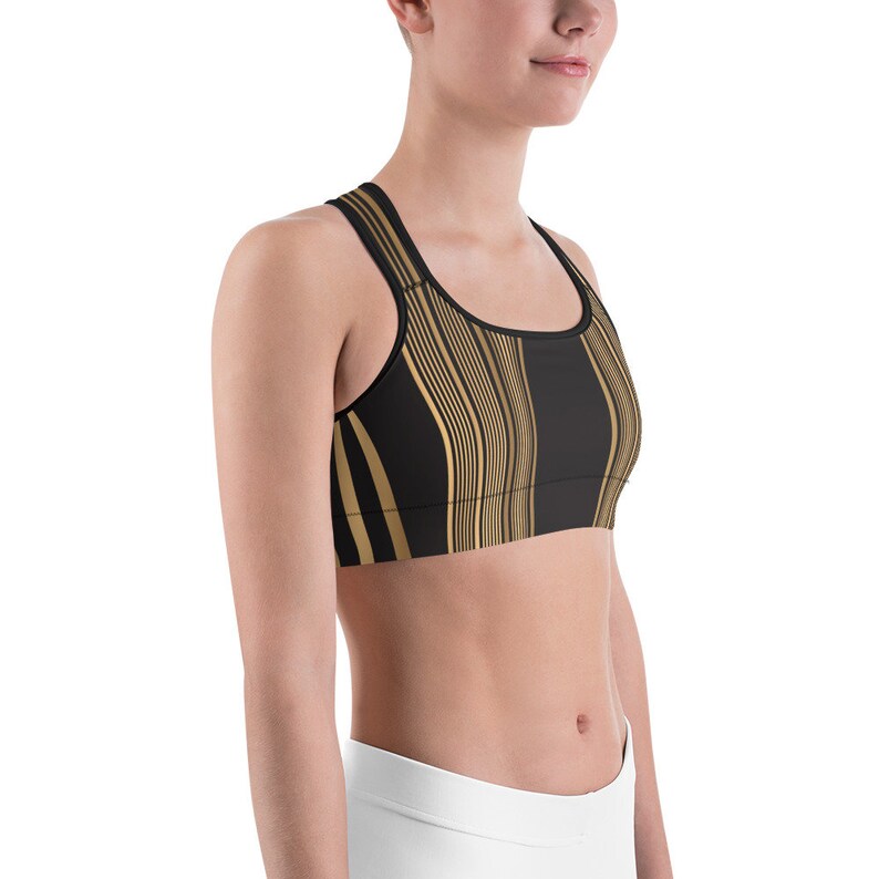 May include: Black sports bra with a gold vertical wavy stripe pattern. The bra has a racerback design and a scoop neckline. The fabric appears to be a soft, stretchy material, suitable for athletic activities.
