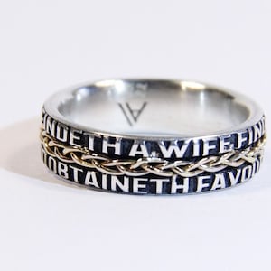 Könnte beinhalten: Ein silberner Ring mit einem goldenen Band und der Inschrift "FINDE THA WIFE FINDE THA WIFE OBTAINETH FAVOUR OBTAINETH FAVOUR" um das Band.