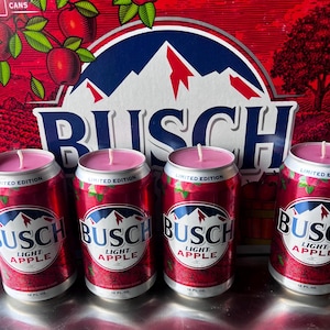 May include: Four red and silver Busch Light Apple beer cans repurposed as candles. Each can has a pink candle top with a white wick. The cans are in front of a red Busch beer box with the Busch logo and apple illustrations.