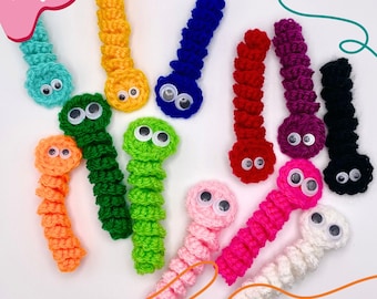 May include: A collection of twelve small, crocheted worm-shaped figures in various colors including turquoise, yellow, blue, red, purple, black, orange, green, light green, pink, hot pink, and white, each featuring two attached plastic googly eyes.