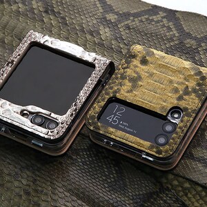 以下が含まれることがあります： 蛇の皮の質感のデザインが施されたスマートフォンケースが2つ。1つのケースは銀色で、黒い画面に「12:45」と「Wed, August 11」の時間が表示されています。もう1つのケースは金色で、黒い画面です。