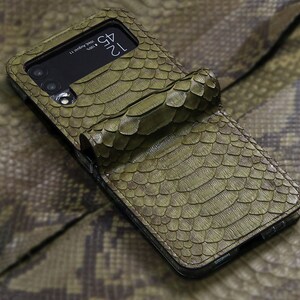 以下が含まれることがあります： カメラレンズを覆うフラップが付いた緑色のヘビ革製携帯電話ケース。ケースはテクスチャ加工された表面と黒い縁取りをしています。