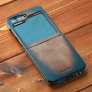 May include: A brown leather phone case with a clear plastic window for the screen. The case has a fold-over flap and the brand name "MOTIMO" is printed on the flap.