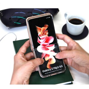 以下が含まれることがあります： クリアケースに入れたローズゴールドのGalaxy Z Flip3 5Gスマートフォンを持った人のクローズアップ。携帯電話の画面には、時刻「12:45」と日付「Wed August 1」が表示されています。