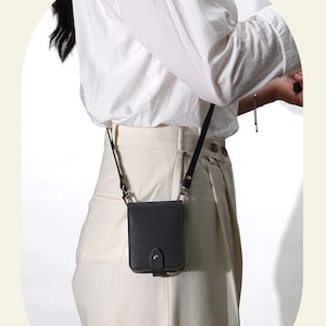 May include: A small, black crossbody bag with a rectangular shape and a flap closure. The bag has a long, thin strap and is made of a textured material. The bag is worn over the shoulder.