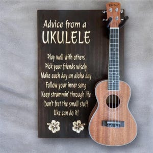 May include: A brown ukulele next to a wooden sign with the text "Advice from a Ukulele." The sign includes inspirational phrases like "Play well with others" and "Don't fret the small stuff." Two white flower designs are at the bottom of the sign.