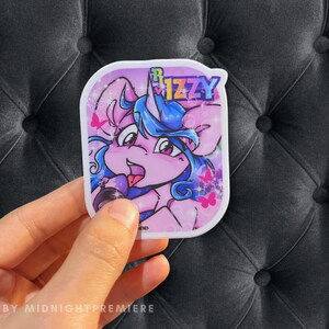 Könnte beinhalten: Ein Aufkleber mit einem Cartoon-Einhorn mit blauen Haaren und rosa Fell, das ein Mikrofon hält. Das Einhorn trägt ein blaues Hemd und hat einen rosa Hintergrund mit Glitzer und Schmetterlingen. Der Text "Rizzy" ist in Regenbogenfarben auf dem Aufkleber geschrieben.