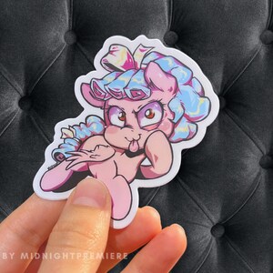 May include: A pink and blue cartoon pony with a blue and white bow on its head. The pony is sticking its tongue out and has a mischievous expression on its face.