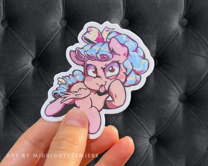 May include: A pink and blue cartoon pony with a blue and white bow on its head. The pony is sticking its tongue out and has a mischievous expression on its face.