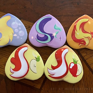 May include: Five heart-shaped pins featuring the ponytails of the main characters from My Little Pony: Friendship is Magic. The pins are yellow, purple, orange, red, and yellow with white.