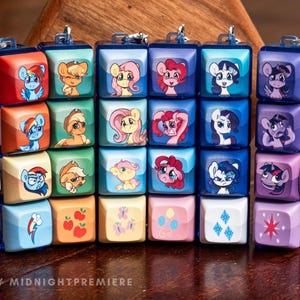 May include: A keychain with small, square blocks, each displaying a different cartoon pony illustration. The blocks are in shades of blue, red, pink, and purple. The keychain is attached to a silver clasp and a dark blue chain.