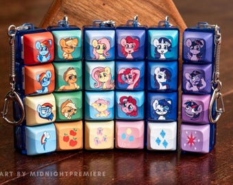 May include: A keychain featuring a series of small, square blocks with cartoon pony illustrations. The blocks are in various colors, including blue, red, pink, and purple. The keychain is attached to a silver clasp and a dark blue chain.