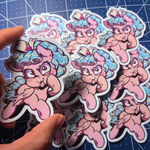 May include: A stack of pink and blue cartoon pony stickers with a tongue sticking out. The stickers have a white background and are printed with a black outline. The stickers are all the same design and are stacked on top of each other. The stickers are on a blue and white checkered surface.