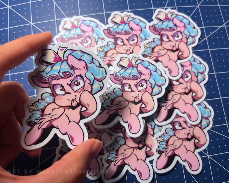 May include: A stack of pink and blue cartoon pony stickers with a tongue sticking out. The stickers have a white background and are printed with a black outline. The stickers are all the same design and are stacked on top of each other. The stickers are on a blue and white checkered surface.