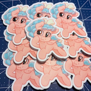 May include: A stack of pink and blue cartoon pony stickers with a white bow. The pony is smiling and has its arm raised in a flexed pose.