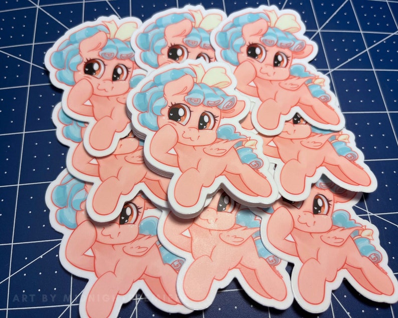 May include: A stack of pink and blue cartoon pony stickers with a white bow. The pony is smiling and has its arm raised in a flexed pose.