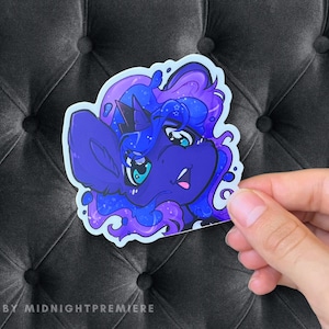 Puede incluir: Una pegatina con un poni de dibujos animados en tonos azules y morados. El poni tiene una melena azul oscuro y un ojo azul claro. La pegatina está sujeta por una mano sobre un fondo negro. El texto "ART BY MIDNIGHTPREMIERE" está en la parte inferior.