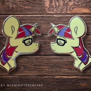 May include: Two gold-coloured enamel pins featuring a cartoon pony with a purple and red mane and a unicorn horn. The pony is wearing glasses and has a yellow body.
