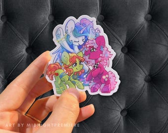 May include: A sticker featuring three cartoon ponies. The pony on the left is green with a red mane and tail. The pony in the middle is blue with a white mane and tail. The pony on the right is pink with a purple mane and tail. All three ponies are wearing colorful outfits and accessories.