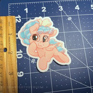 May include: A pink pony with blue and white hair, wearing a yellow bow, is depicted in a cartoon style. The pony is smiling and has its head tilted to the side. The background is a blue and white grid.