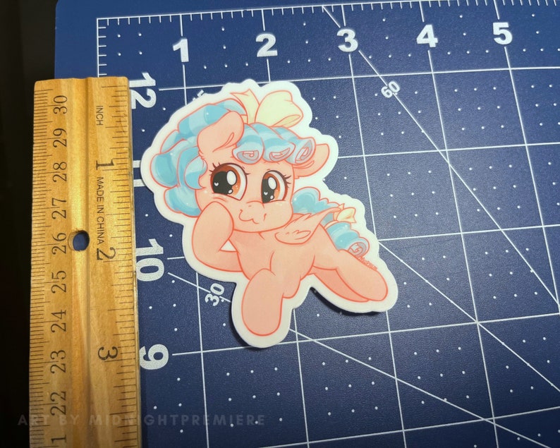May include: A pink pony with blue and white hair, wearing a yellow bow, is depicted in a cartoon style. The pony is smiling and has its head tilted to the side. The background is a blue and white grid.