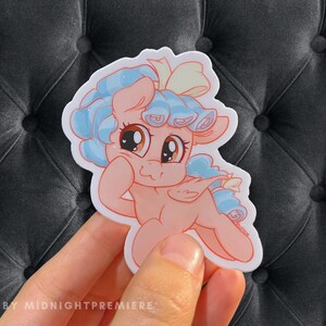 May include: A sticker featuring a cartoon pony with light blue hair, a pink body, and a white bow. The pony has large, brown eyes and a surprised expression. The sticker is on a gray background.