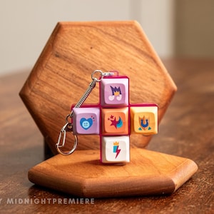 Könnte beinhalten: Ein rosa-weißer Schlüsselanhänger mit sechs bunten Tasten, die verschiedene Symbole zeigen. Die Tasten sind kreuzförmig angeordnet und an einer silbernen Kette befestigt.