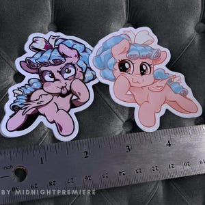 May include: Two pastel pink pony stickers with blue hair and bows. The pony on the left is sticking out its tongue. The pony on the right is resting its head on its hand. The stickers are on a gray fabric background with a ruler underneath.