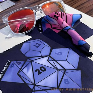 Peut inclure: Deux chiffons de nettoyage en microfibre sur le thème des dés. Un chiffon est bleu avec un grand dé à 20 faces et le texte "TRUST THE DICE". L'autre chiffon est rose et bleu avec un dé à 20 faces et le texte "THE DICE".