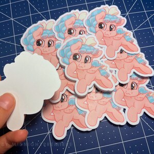 May include: A stack of pink and blue cartoon pony stickers with a white bow on their heads. The ponies are in a sitting position with their legs crossed and their arms crossed. The stickers are on a blue and white grid background.