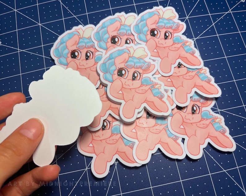 May include: A stack of pink and blue cartoon pony stickers with a white bow on their heads. The ponies are in a sitting position with their legs crossed and their arms crossed. The stickers are on a blue and white grid background.