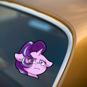 May include: A cartoon sticker of a purple pony with a worried expression. The pony has a purple mane with blue and white streaks, a white horn, and large, expressive eyes. The sticker is placed on a car window.