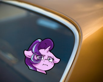 Può includere: Un adesivo a fumetti di un pony viola con un'espressione preoccupata. Il pony ha una criniera viola con striature blu e bianche, un corno bianco e grandi occhi espressivi. L'adesivo è posto sul finestrino di un'auto.
