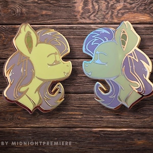 May include: Two enamel pins depicting cartoon horses with purple and green hair. The horses are facing each other with their eyes closed. The pins are displayed on a wooden surface.