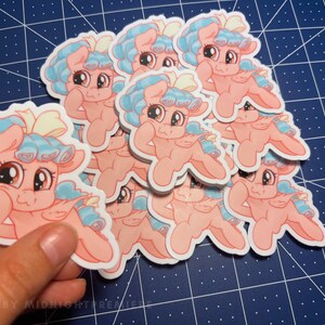 May include: A stack of pink and blue cartoon pony stickers with a yellow bow. The pony has a light blue mane and tail. The stickers are on a blue grid background.
