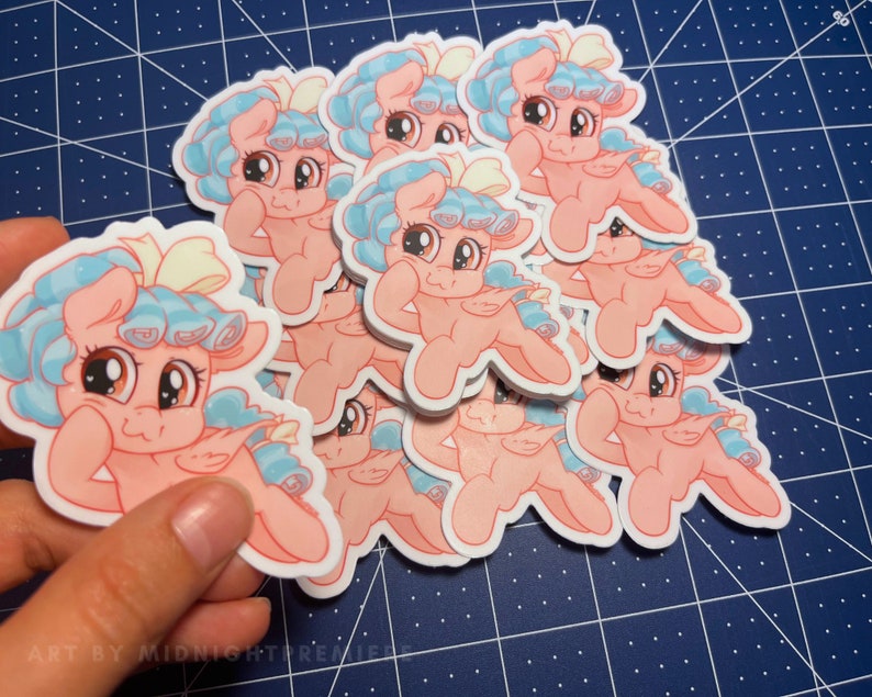 May include: A stack of pink and blue cartoon pony stickers with a yellow bow. The pony has a light blue mane and tail. The stickers are on a blue grid background.