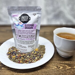 May include: A packet of "i NEED GOURMET SLEEPING DRAGON" herbal tea is on a white plate with loose tea leaves. A teacup with tea is on the right. The tea is caffeine-free.