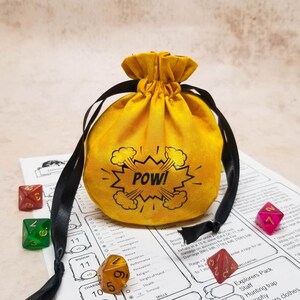 Small yellow dice bag with POW in a superhero comic book style speech bubble. Bag is closed and sits on a character sheet with some dice