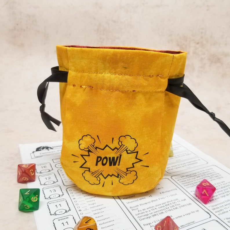 Small yellow dice bag with POW in a superhero comic book style speech bubble. Bag is open and sits on a character sheet with some dice
