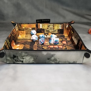May include: A square dice tray with a detailed interior scene, featuring dice. The tray has a grey exterior with a landscape design and black snap closures. Several multi-sided dice are inside the tray, with additional dice visible.