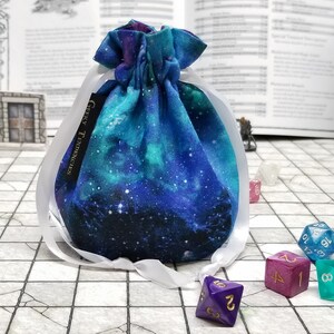 May include: A blue and green galaxy print drawstring bag with white ribbon ties. The bag is filled with dice, including a purple d20, a pink d6, a blue d8, a green d5, and a blue d12. The bag has a label that reads "Geeky Trinkets".