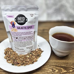 May include: A bag of "i NEED GOURMET GALACTIC SPICE" herbal tea, a cup of tea, and a plate of loose tea leaves. The bag has a purple label with a cartoon image and the words "Caffeine Free".